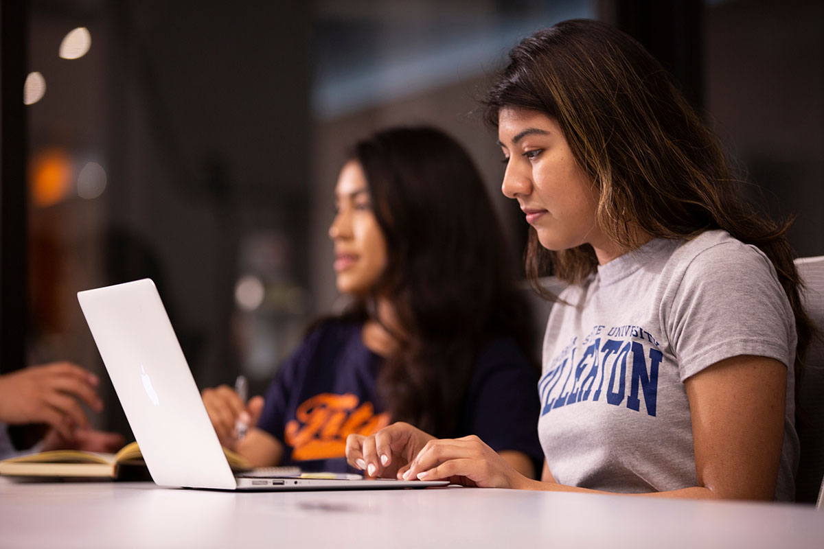 Two Cal State Fullerton students sitting in a library, working on their laptops.