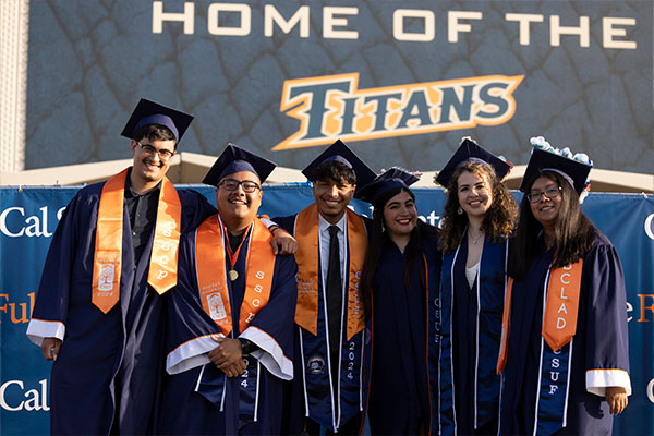 Six Cal State Fullerton students, three males and three females, stand shoulder to shoulder in graduation gowns