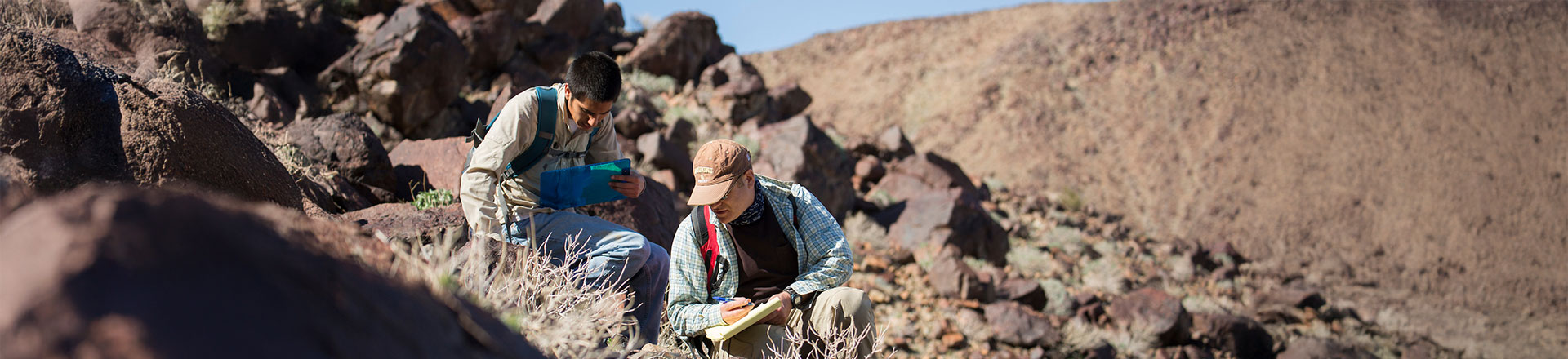 Two researchers in outdoor gear sitting among desert rocks and taking notes on clipboards during a field study.